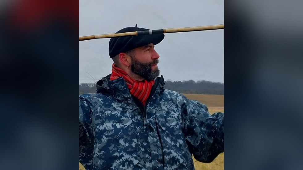 Тук виждаме мъж с брада, който носи барета, оранжев шал и синьо водоустойчиво яке.  Той държи копие, готов да го хвърли.