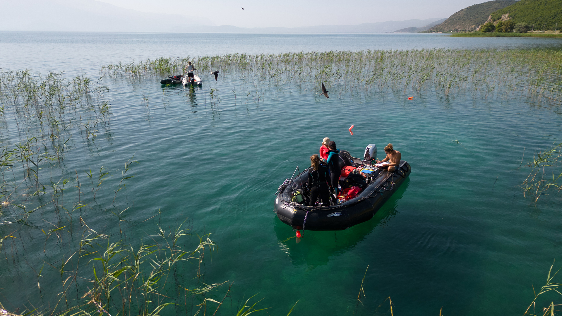 Виждаме надуваема черна лодка с изследователи в нея близо до брега на езеро.