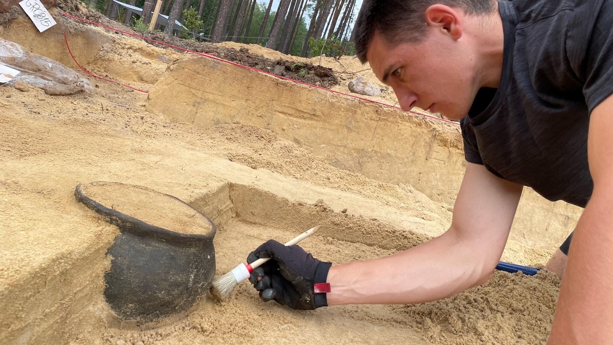 Изследовател изкопава черна урна за кремация в пълно с мръсотия пространство за разкопки.