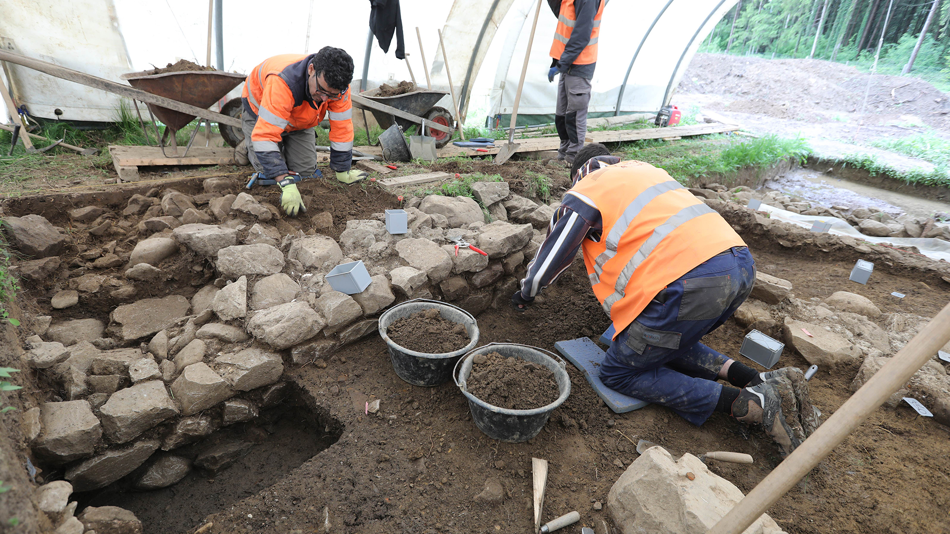 Археолози в якета с висока видимост разкопават ниска стена.