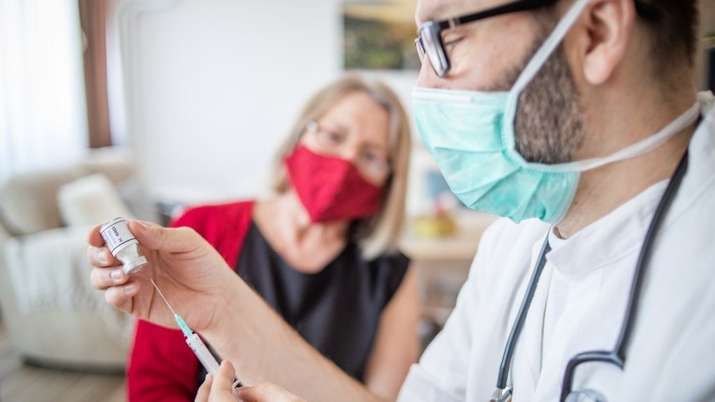 Мъж с хирургическа маска и ръкавици подготвя ваксина срещу COVID-19 за маскиран пациент, който седи на заден план