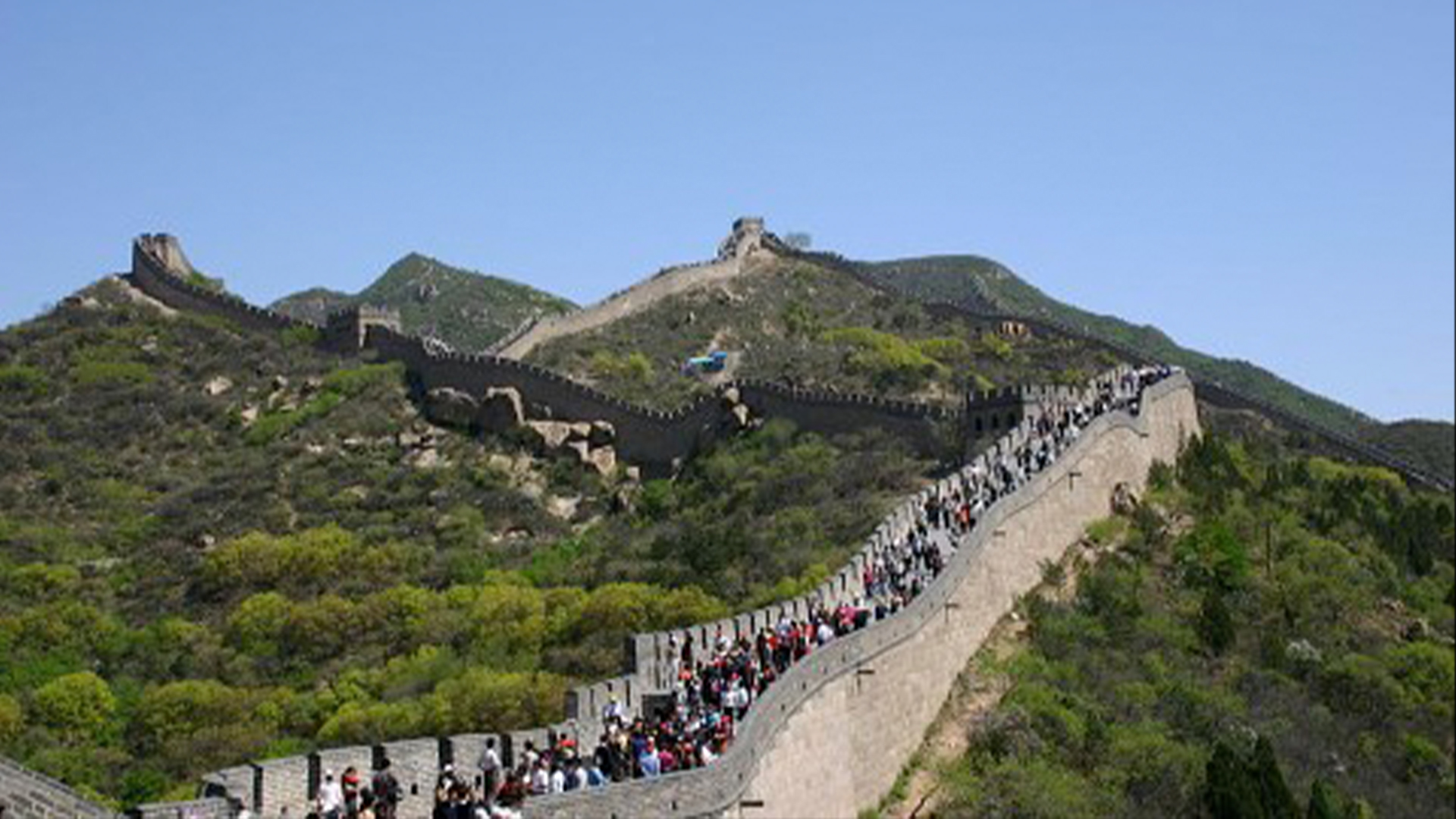 Голям участък от Великата китайска стена в Бадалинг, пълен с хора, докато се разхождат по билото.  Великата стена пресича зелена планинска верига под синьо небе.