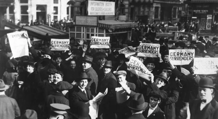 A crowd of men hold up newspapers with the headline "Germany surrenders"