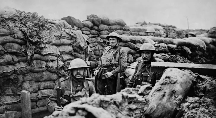 Three men sit in a trench with their helmets and weapons