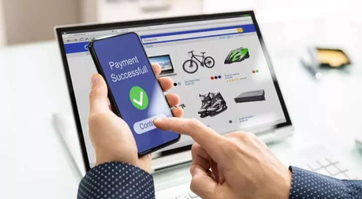 Лаптоп показва онлайн продукти, налични за покупка, докато човек държи телефон, показващ „Успешно плащане“
