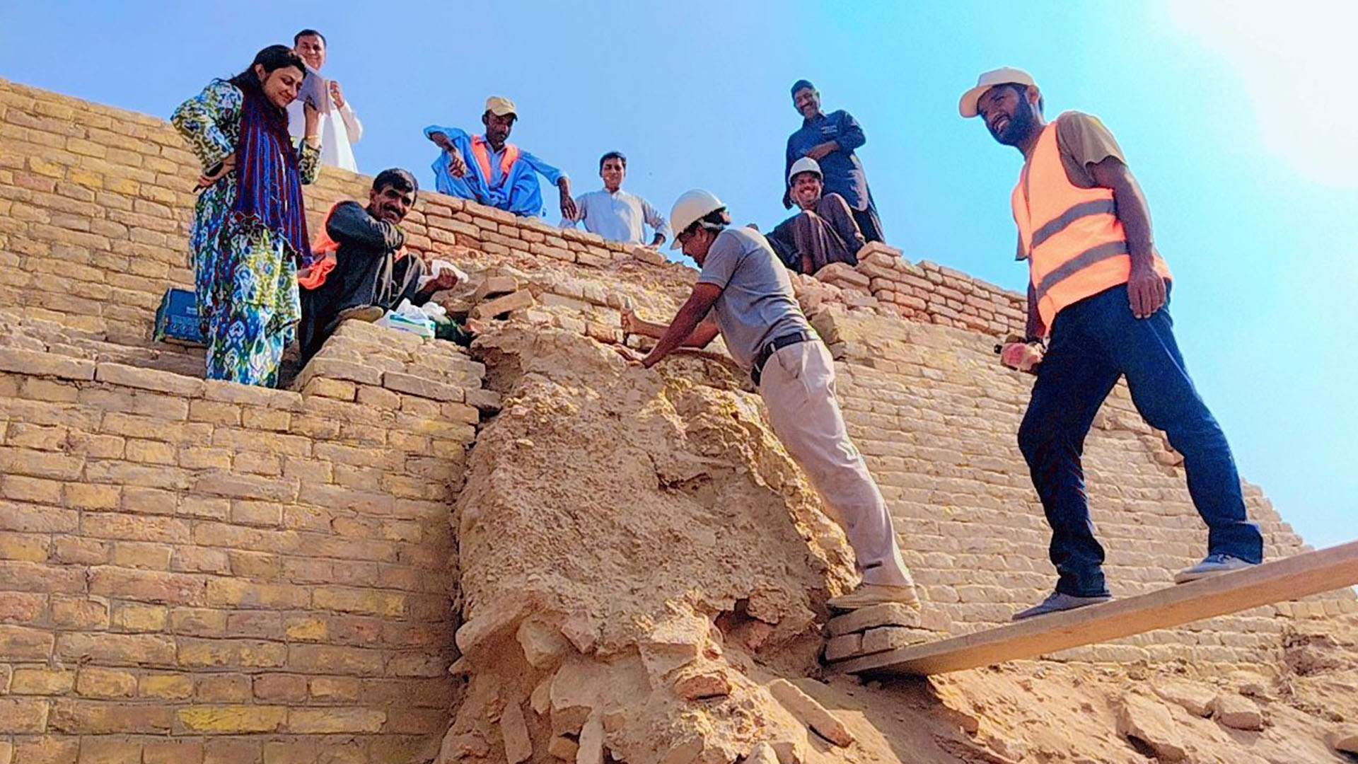 Екип от девет археолози и изследователи стои върху руините на будистко светилище, направено от жълти тухли и пясък.