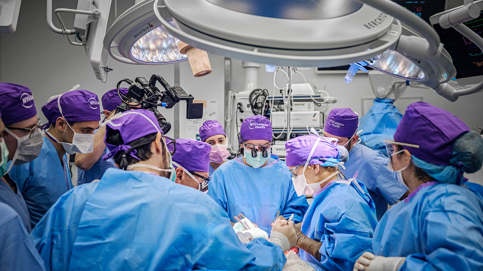 Лекари в сини медицински престилки и лилави хирургически шапки извършват операция в операционна зала.