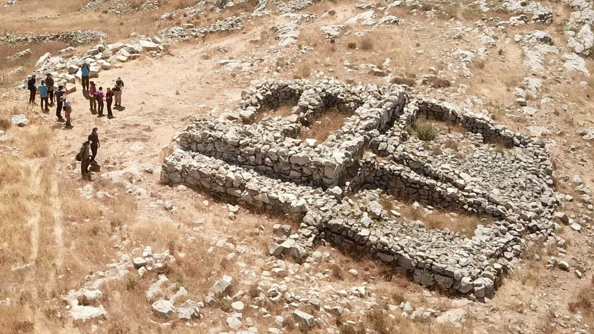 Купчина каменни отломки върху място за археологически разкопки върху кафява пръст.  Група изследователи стои отляво.