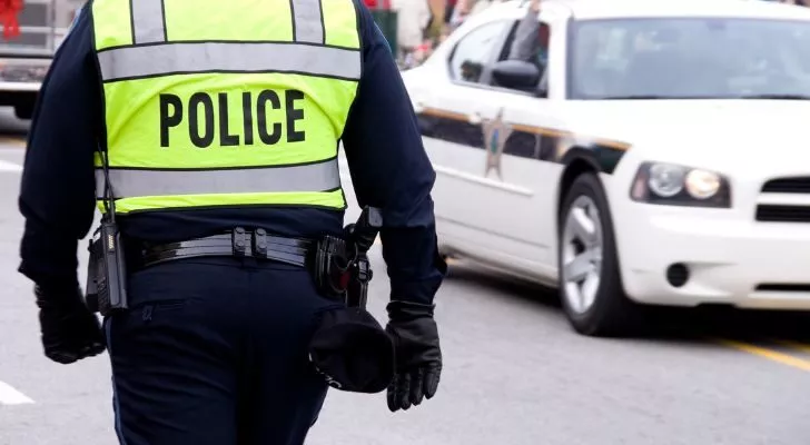 Мъж в полицейска униформа с високо видимо яке с надпис „полиция“ върви по улица