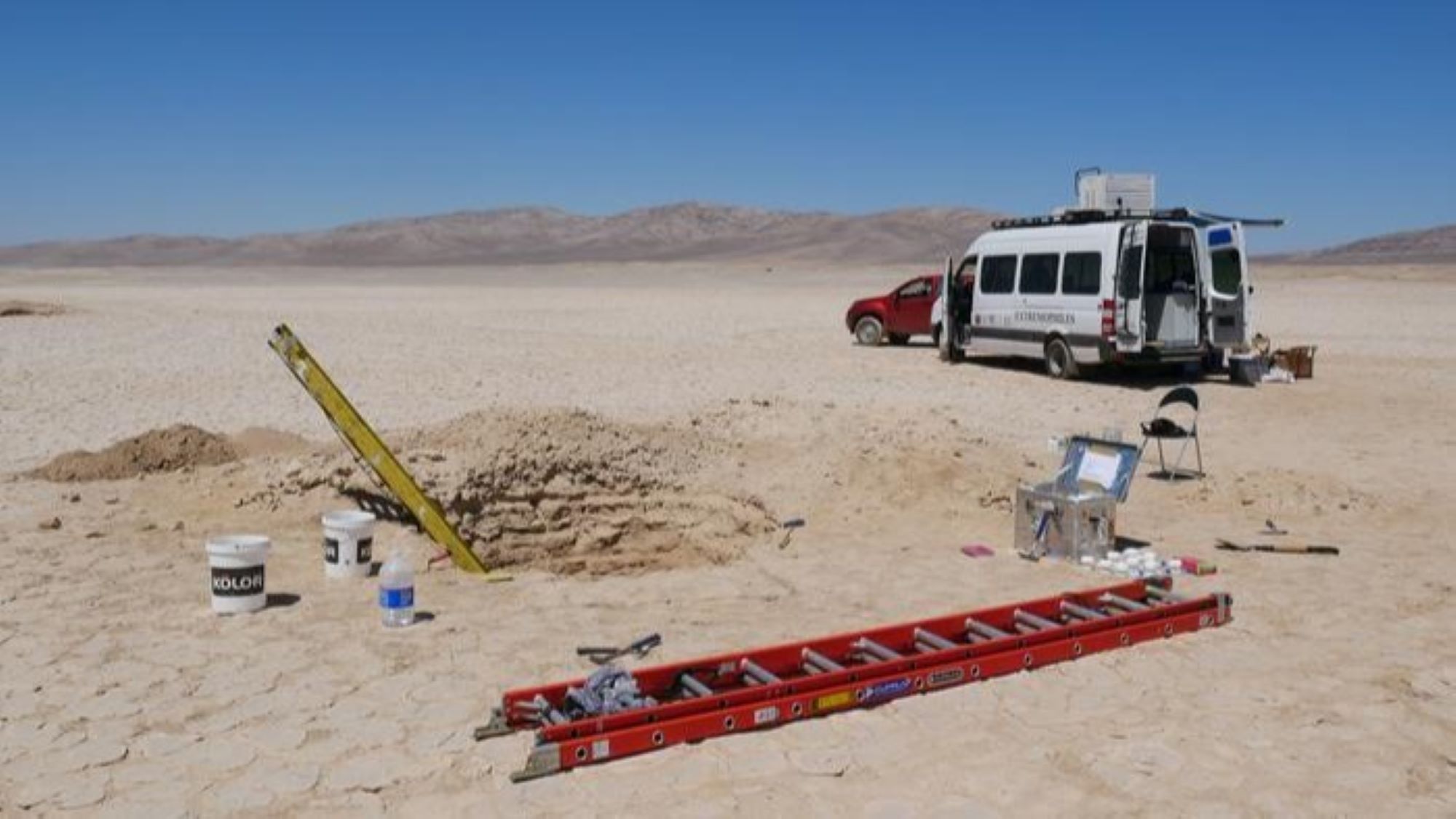 Мястото за изследване в пустинята Атакама, изобразено с камион и микробус на заден план и стълби и инструменти на преден план