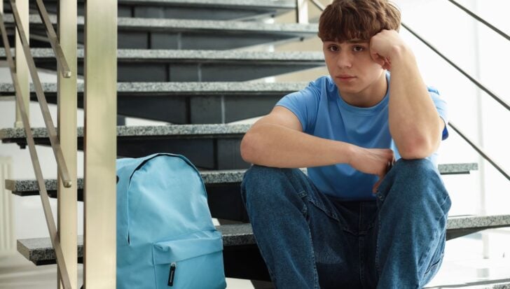 Млад човек, седнал на стълби, изглеждащ замислен със синя раница наблизо