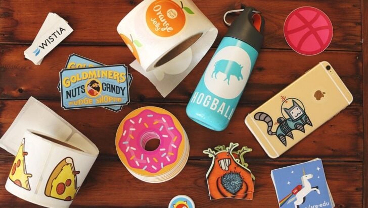 Различни стикери, показани на маса, включително дизайни на поничка, парче пица и герой в космически костюм. Виждат се също бутилка вода, телефон със стикер и ролки стикери.